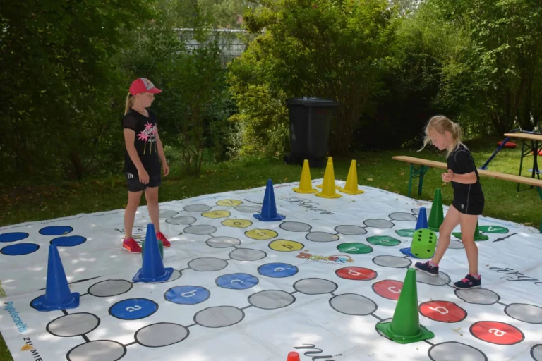 Zwei Mädchen spielen ein Brettspiel im Freien mit farbigen Kegeln und einem großen Würfel.