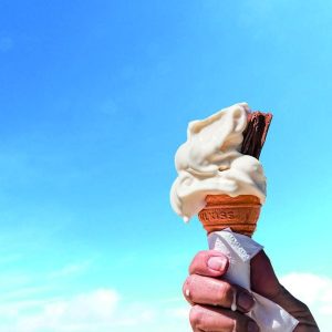 Hand hält ein Eis mit Schokoladenriegel vor blauem Himmel.