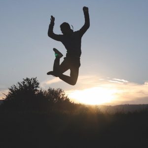 Eine Person befindet sich mitten im Sprung im Freien, die Silhouette hebt sich von einem Sonnenuntergangshimmel mit Bäumen im Hintergrund ab.