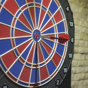 Ein rot-schwarzer Dartpfeil steckt im Bullseye einer blau-rot-weißen Dartscheibe, die an einer Steinwand befestigt ist.