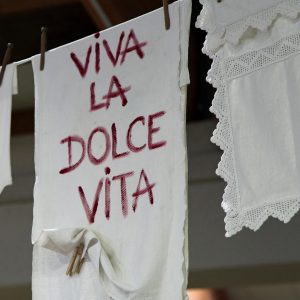 Ein weißes Tuch mit dem rot aufgesprühten Schriftzug Viva la Dolce Vita" hängt an einer Wäscheleine neben spitzenbesetzter Wäsche, die von hölzernen Wäscheklammern gehalten wird.