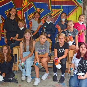 Gruppe von Jugendlichen und Erwachsenen mit Farbsprühdosen vor einem Graffiti-Hintergrund während eines Graffiti-Workshops.