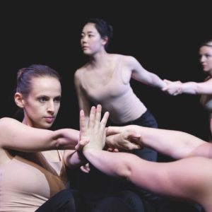 Vier Frauen in farblich neutralen Oberteilen führen vor einem dunklen Hintergrund eine zeitgenössische Tanznummer auf, bei der sie sich gegenseitig an den Armen halten und balancieren.
