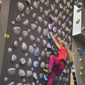 Ein junges Mädchen in Rosa klettert an einer Boulderwand mit beleuchteten Klettergriffen.