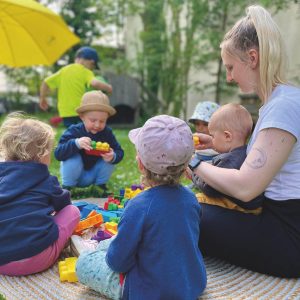 Mehrere kleine Kinder und ein Erwachsener sitzen auf einer runden Matte im Freien und spielen mit bunten Bauklötzen auf einer Wiese unter Bäumen.