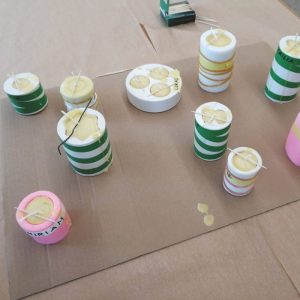 Mehrere zylindrische Behälter, teilweise mit Namen beschriftet, sind mit einer gelblichen Substanz gefüllt und auf einem großen Stück Pappe angeordnet.