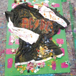 Ein schwarzes AC/DC-Shirt mit weißen Ärmeln ist auf einer grünen Tafel ausgestellt, die mit Glitter, Konfetti und verschiedenen kleinen Objekten wie Perlen, Kacheln und Sternformen bedeckt ist.