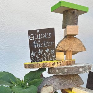 Eine handgefertigte Holzskulptur mit einem kleinen Schild mit der Aufschrift "Glücks Wächter" und einfachen Blumenzeichnungen, platziert neben einer Grünpflanze vor einem weißen Wandhintergrund.