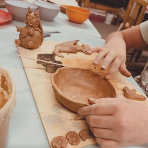 Eine Person formt eine Tonschale auf einem Holzbrett, wobei kleine Tonfiguren, Blumen und Werkzeuge um ihre Hände herum sichtbar sind.