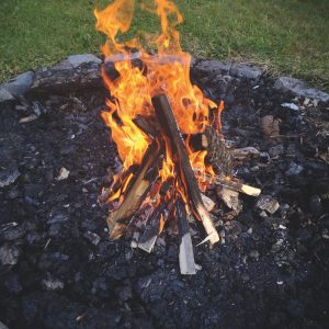 Ein Lagerfeuer mit brennenden Holzscheiten und Flammen in einem Steinfeuerring, umgeben von Gras und einem Stuhl im Hintergrund.