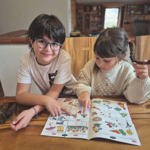 Ein Junge und ein Mädchen sitzen an einem Holztisch und schauen auf ein aufgeschlagenes Activity-Buch mit bunten Illustrationen und Rätseln.