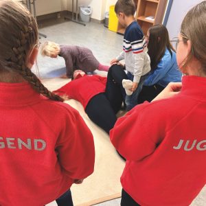 Kinder üben Erste-Hilfe-Maßnahmen an einer am Boden liegenden Person; zwei tragen rote Jacken mit der Aufschrift JUGEND auf dem Rücken.