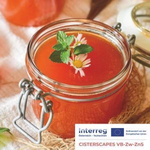 Ein mit Orangenmarmelade oder -gelee gefülltes Glas, garniert mit Minzblättern und kleinen Blumen, auf einem Tuch platziert. Die Logos der EU und des Interreg-Projekts sind in der unteren rechten Ecke zu sehen.