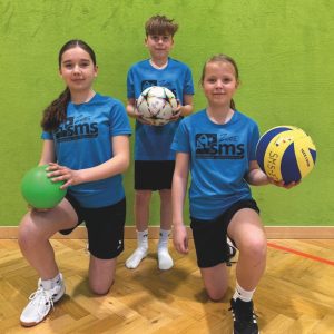 Drei Kinder in blauen Hemden und schwarzen Shorts posieren in einer Turnhalle und halten jeweils einen anderen Ball (grün, Fußball und Volleyball) gegen eine grüne Wand.