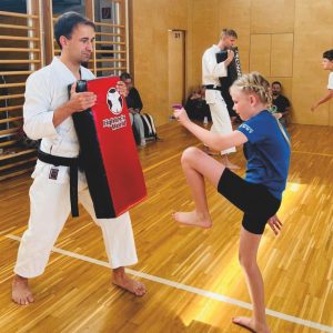 Ein Kind in Sportkleidung übt einen Kniestoß auf ein gepolstertes Schild, das von einem Lehrer in einer Turnhalle mit Holzböden und -wänden gehalten wird. Andere Personen beobachten im Hintergrund.
