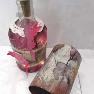 Dekorative Glasflasche mit braunem Korken, verziert mit Spitze, rosafarbenen Papierfeen und einem lilafarbenen, blumengemusterten Etikett; daneben steht ein passender zylindrischer Behälter.