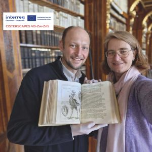 Zwei Personen stehen in einer Bibliothek, lächeln in die Kamera und halten einen alten Bildband aufgeschlagen; im Hintergrund sind Bücherregale und ein Projektlogo zu sehen.
