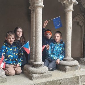 Vier Kinder und eine junge Frau sitzen hinter Steinsäulen und halten die Flaggen der Europäischen Union und der Tschechischen Republik.