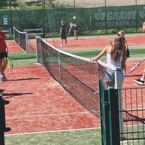 Kinder spielen Tennis auf Sandplätzen im Freien, einige stehen am Netz, andere halten Schläger, während ein Tennisball in der Luft liegt.