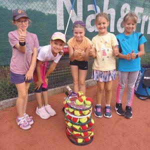 Fünf junge Mädchen auf einem Tennisplatz lächeln und zeigen die Daumen nach oben hinter einem hohen Stapel von Tenniskörben, die mit Bällen gefüllt sind.