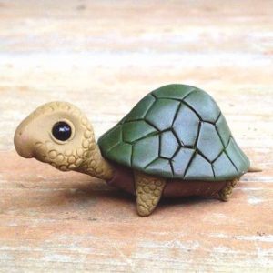 Eine kleine, handgefertigte Schildkrötenfigur mit grünem Panzer und braunem, strukturiertem Körper auf einer Holzoberfläche.
